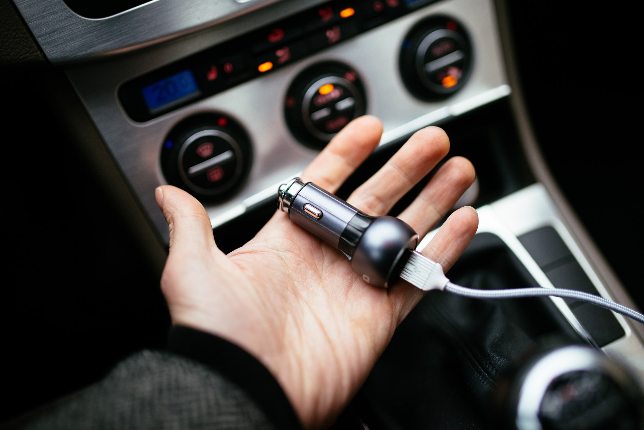 A hand holding a USB car charger in front of a 12V power outlet.
